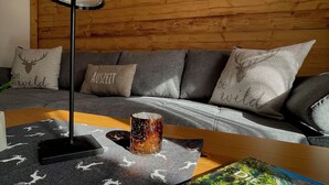 A modern living room with a wooden wall, a grey sofa with patterned cushions, a black floor lamp, a decorative rug with deer prints, and a framed picture of a mountain landscape. - Ferienwohnung, 1-2 Personen, 86 qm, Separates Schlafzimmer, Küche, Wohnzimmer, Du/wc, Balkon (Schönau am Königssee)