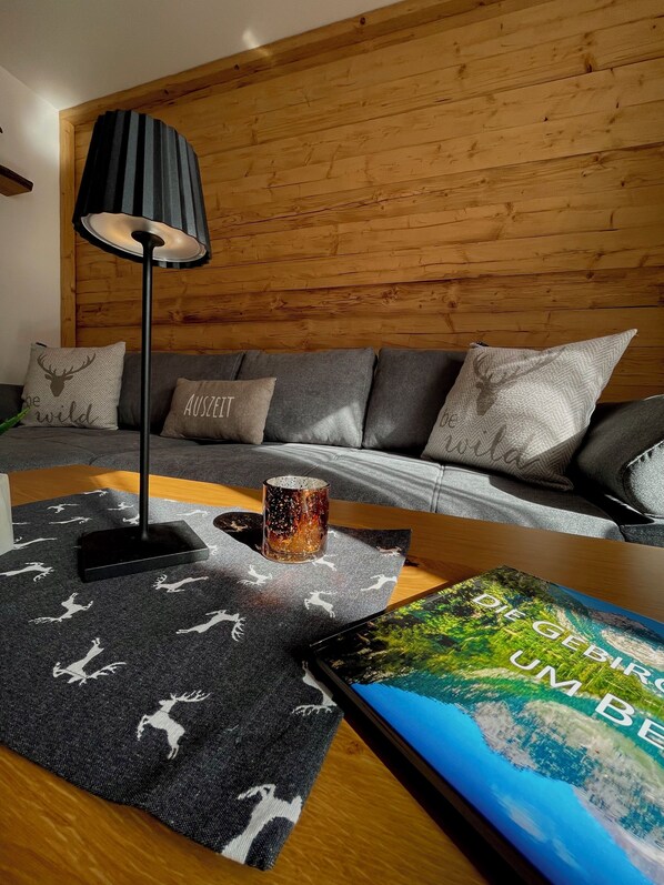 Una sala de estar moderna con una pared de madera, un sofá gris con cojines estampados, una lámpara de pie negra, una alfombra decorativa con estampados de ciervos y un cuadro enmarcado con un paisaje de montaña.