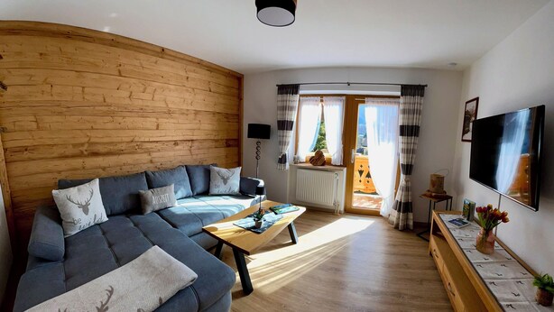 A cozy living room with a wooden wall, a grey sofa, a wooden coffee table, and a TV on the wall. - Ferienwohnung, 1-2 Personen, 86 qm, Separates Schlafzimmer, Küche, Wohnzimmer, Du/wc, Balkon (Schönau am Königssee)