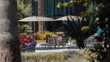 Un patio al aire libre con mesas y sillas, rodeado de plantas y una palmera grande.