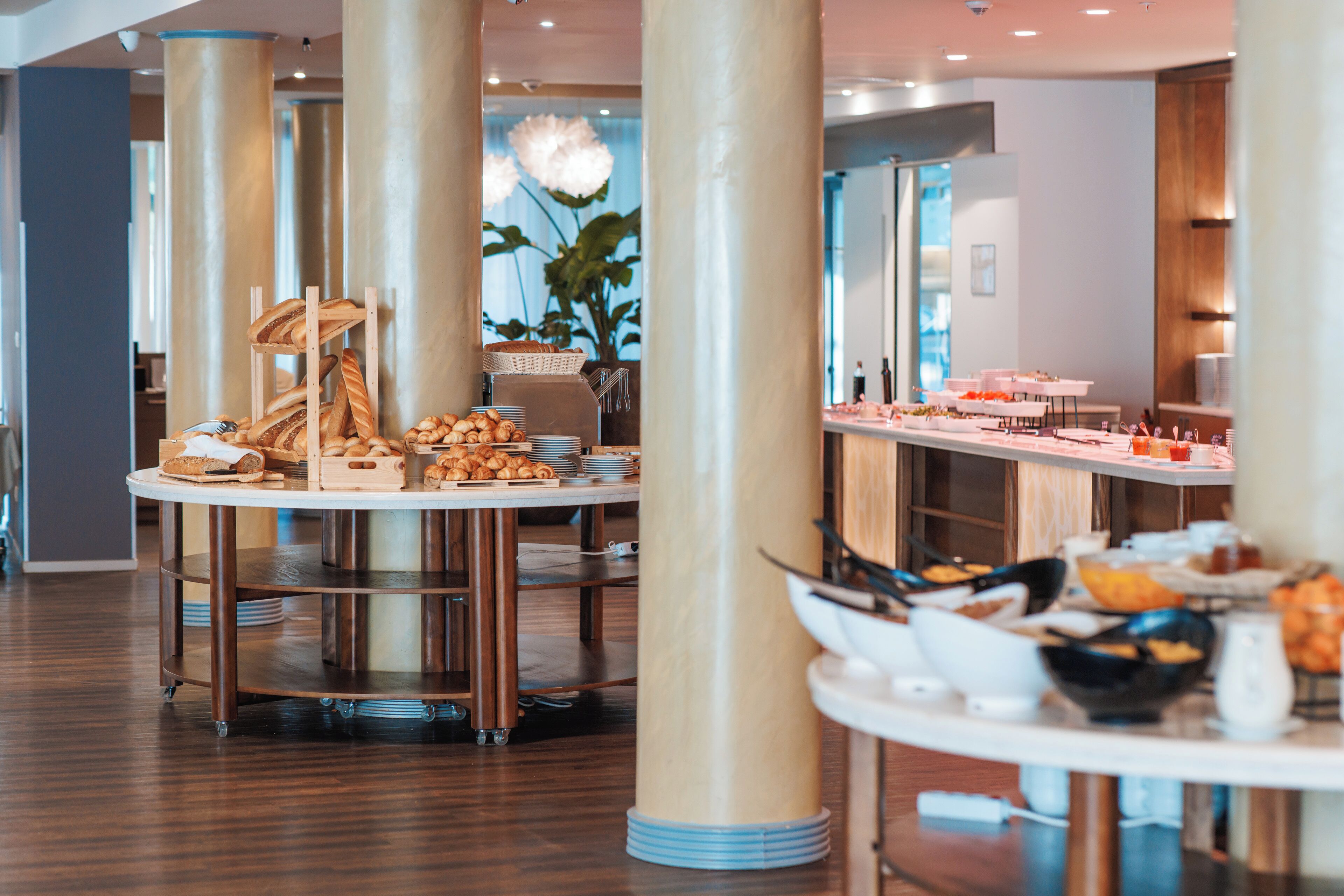 A modern dining area with round tables, a variety of breads and pastries, and a large plant in the background.