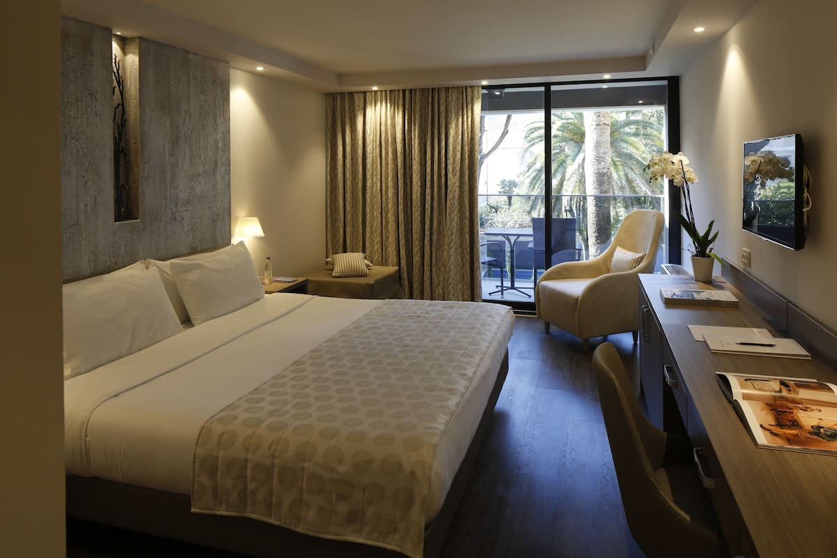 A hotel room with a large bed, a desk with a chair, a TV, and a balcony with a view of palm trees.