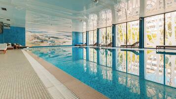 An indoor swimming pool with clear glass walls, lounge chairs, and a tiled floor.