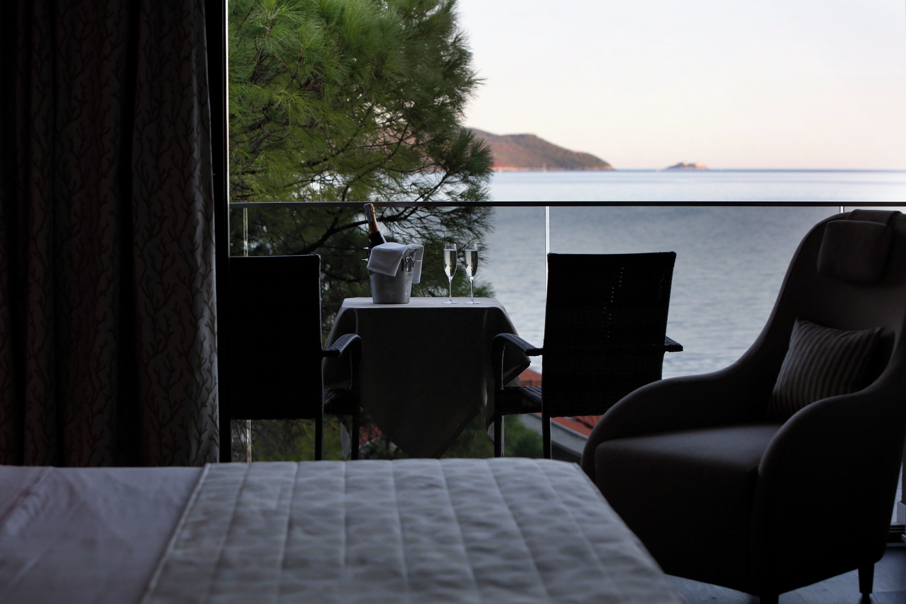 A hotel room with a bed, a table with a bottle and glasses, and a chair facing a balcony with a view of the sea and trees.