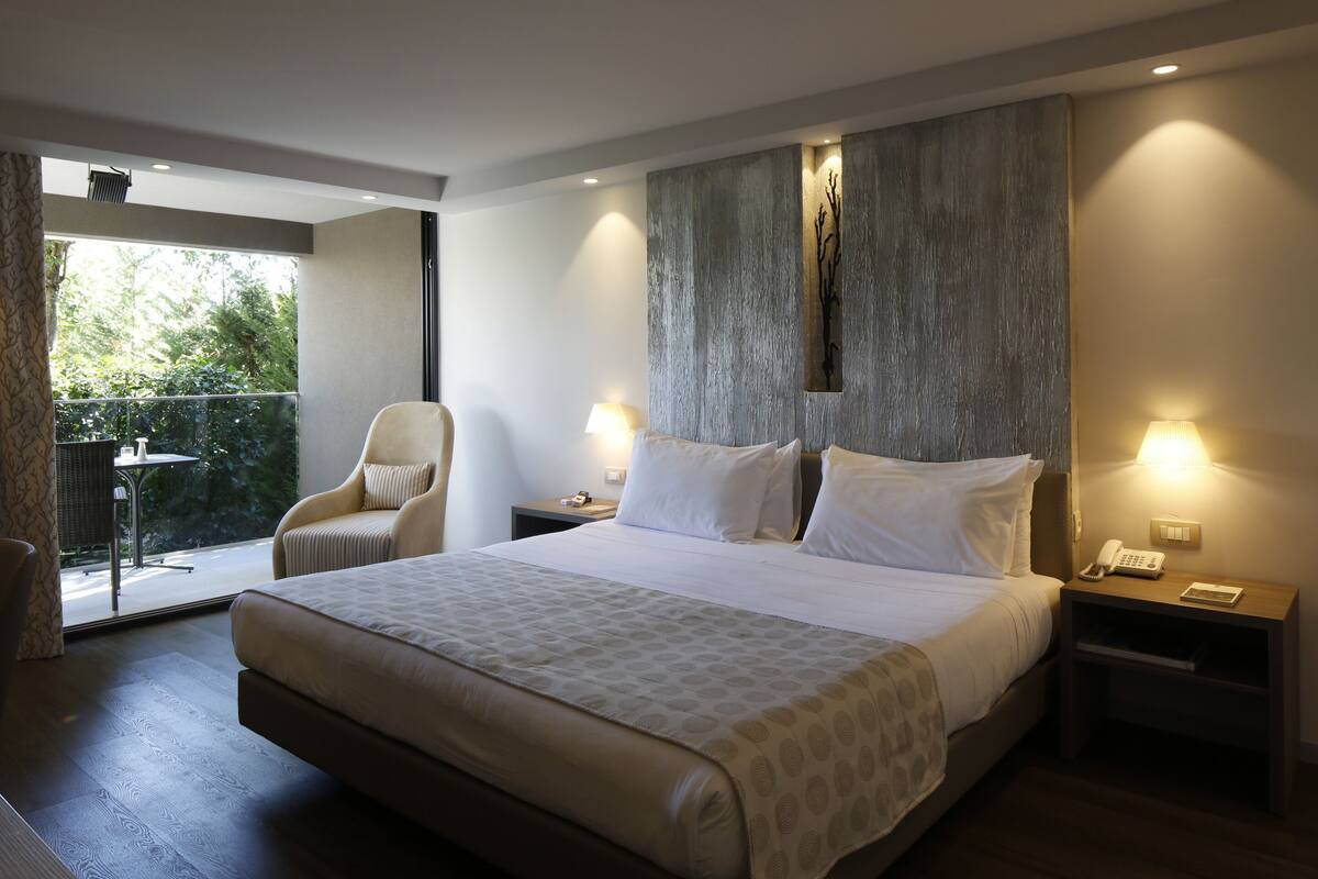 A modern hotel room with a large bed, a chair, a small table, and a balcony with a view of greenery.