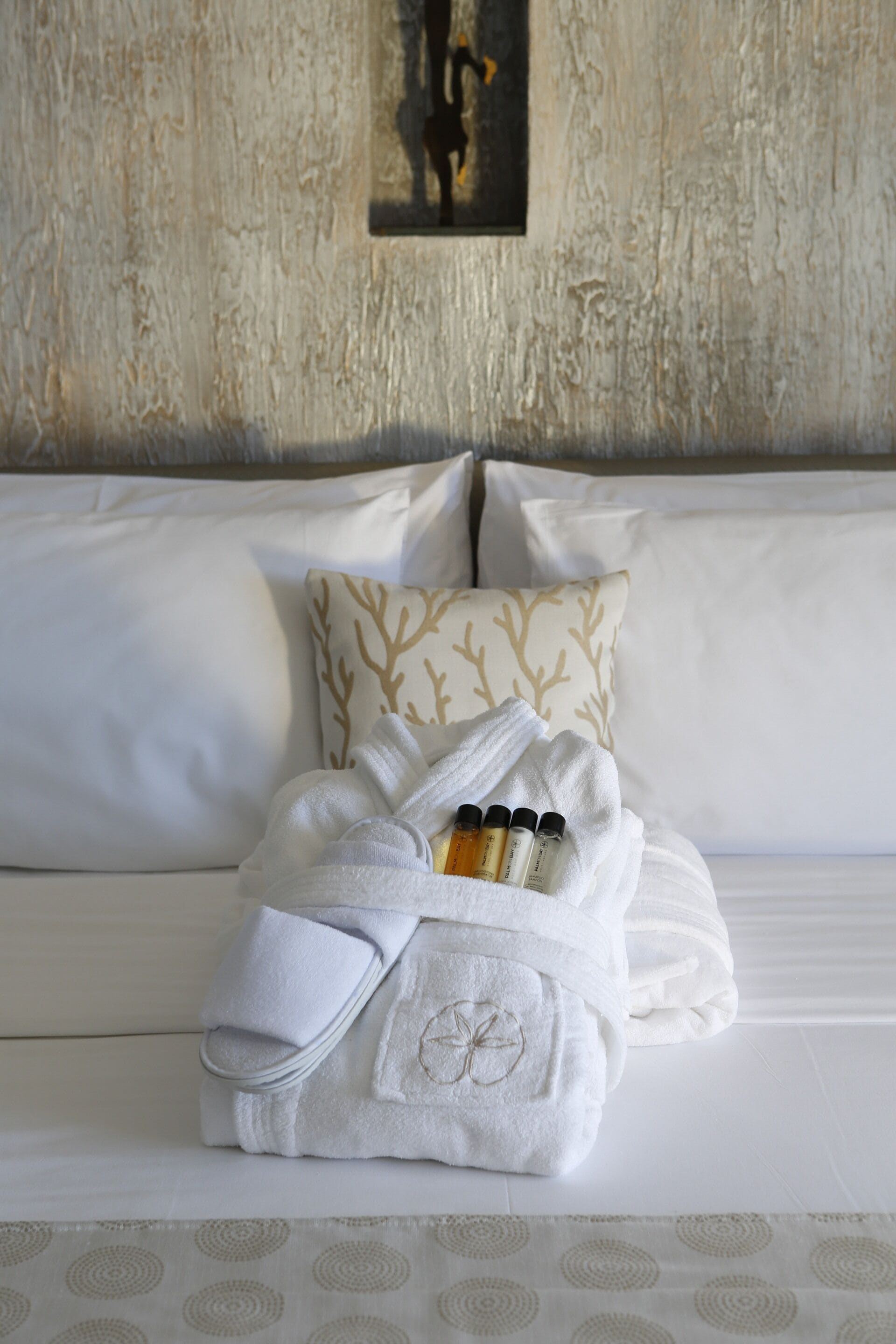 Una cama bien hecha con ropa blanca, un cojín decorativo con estampado de árbol y una toalla con diseño floral.