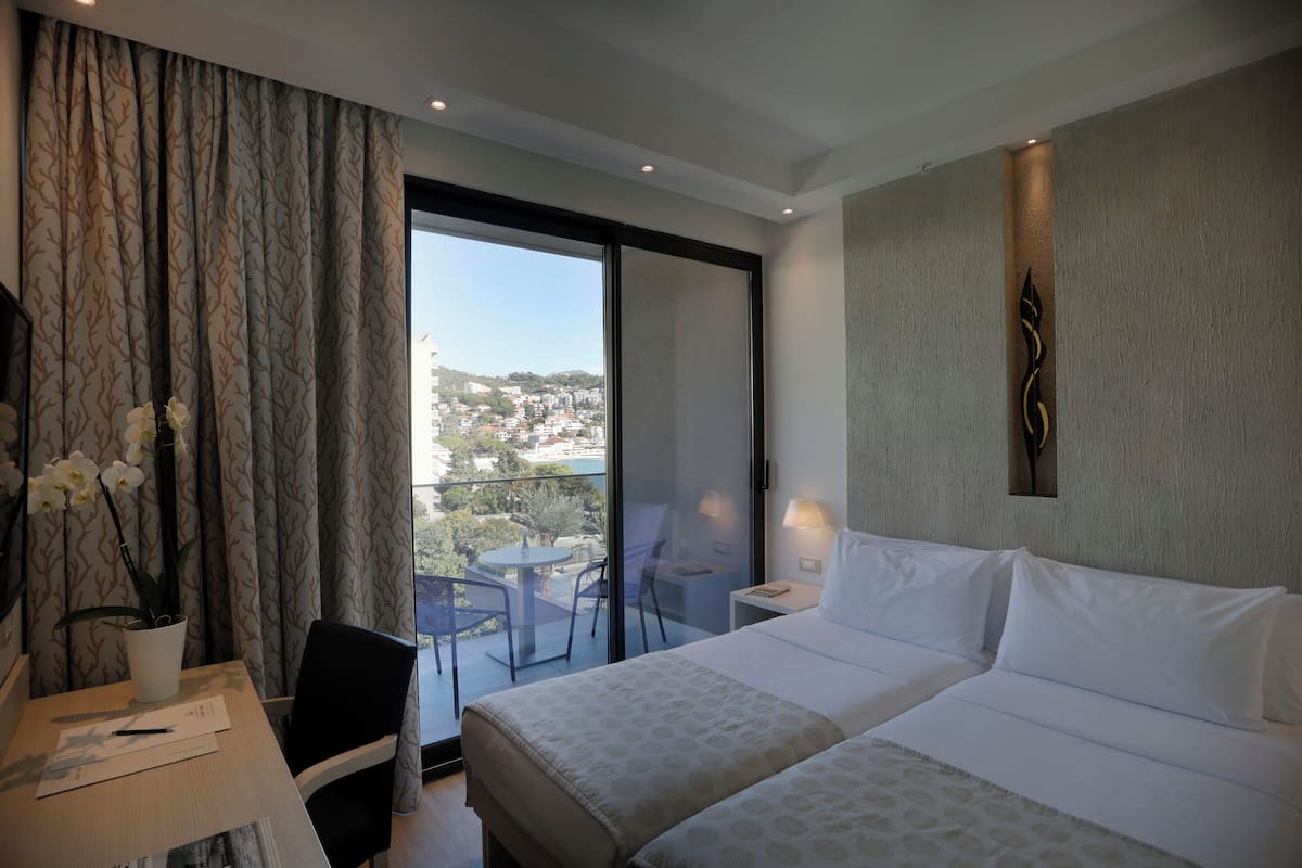 A hotel room with a bed, a desk, a chair, a balcony with a view of the city, and a decorative wall piece.