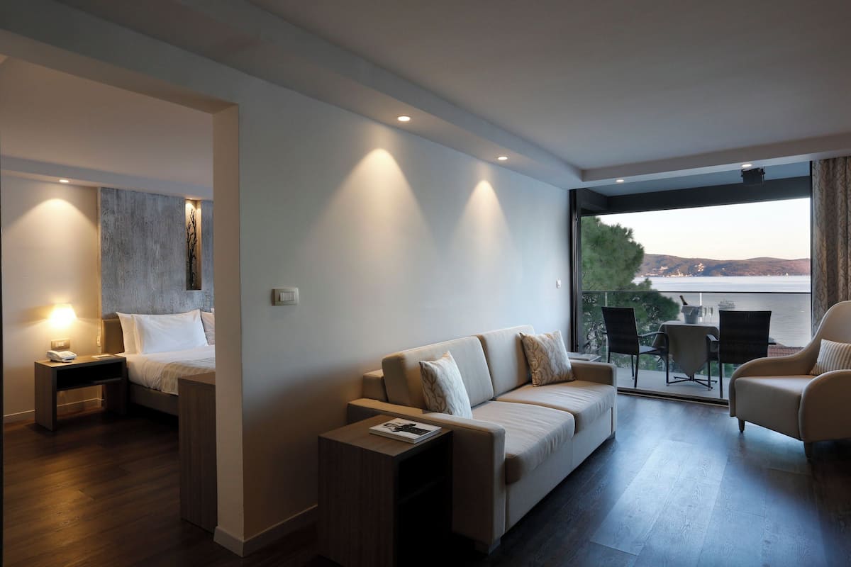 A modern hotel room with a bed, a sofa, a small table, and a balcony with a view of the water.