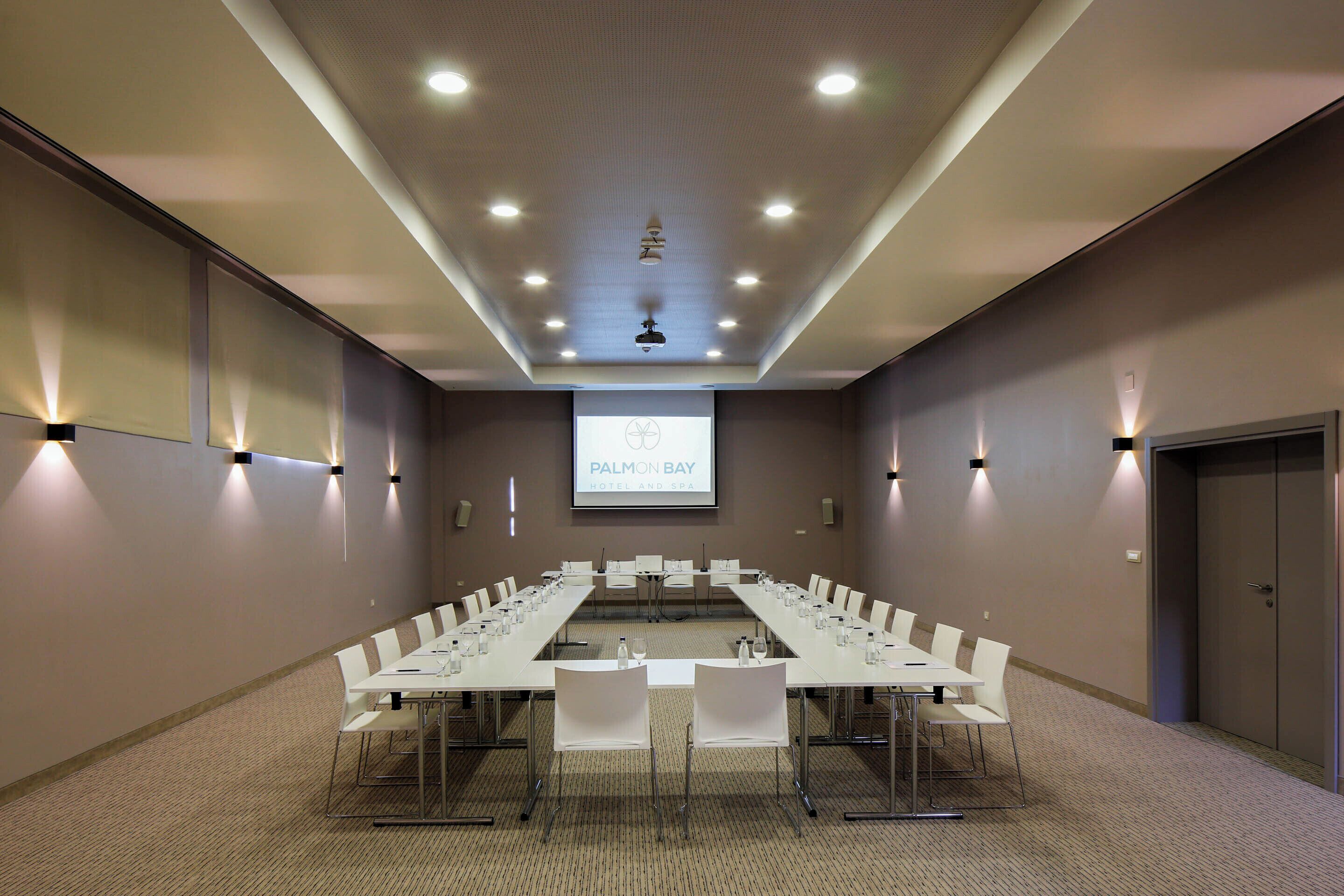 Un salón de conferencias con una larga mesa, sillas y una pantalla proyectora que muestra 