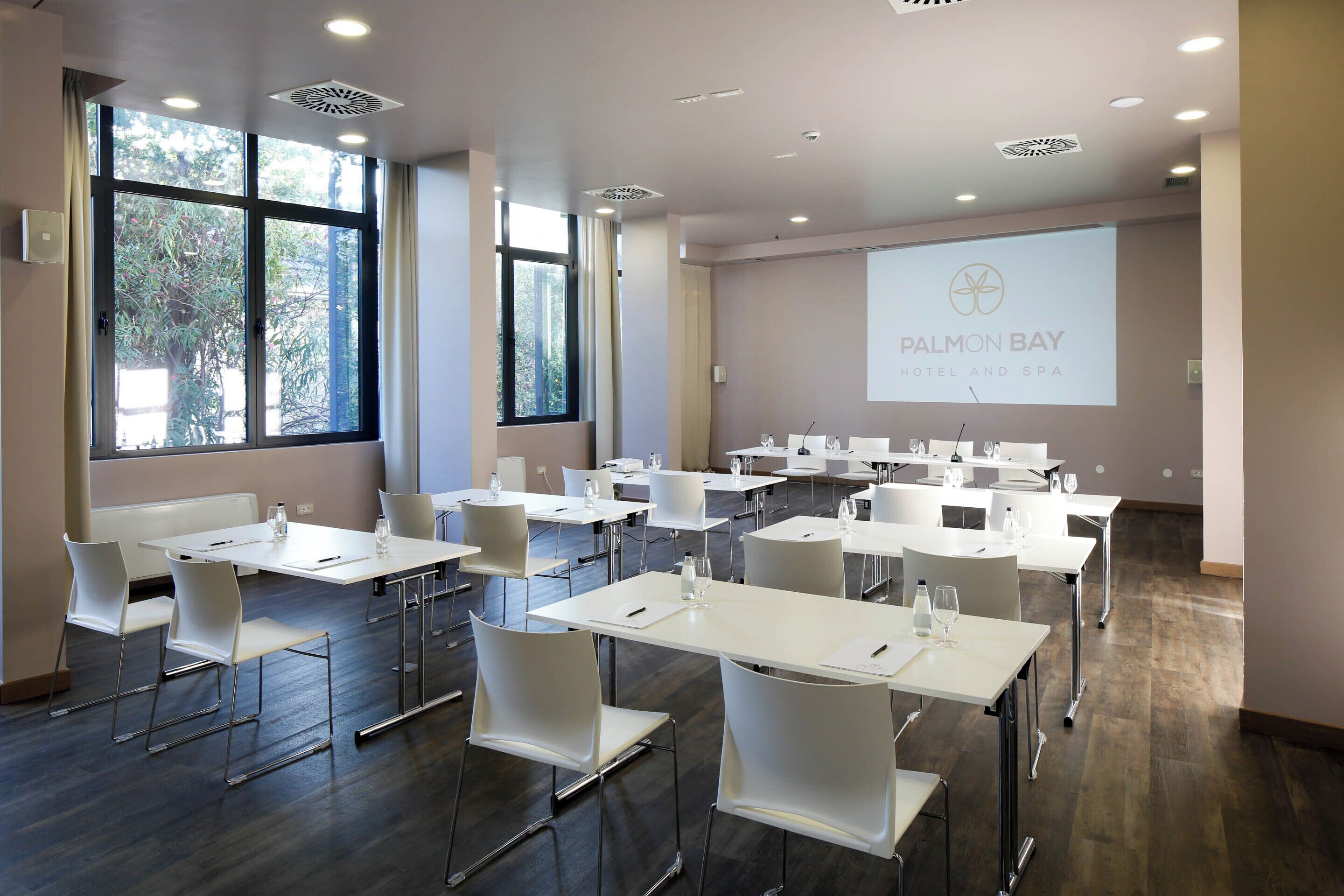 Une salle de conférence avec des tables et des chaises blanches, de grandes fenêtres et un écran de présentation affichant le texte « PALMON BAY HOTEL AND SPA ».