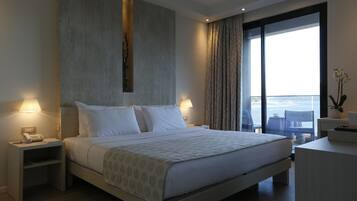 A modern hotel room with a large bed, bedside lamps, a nightstand, a desk, and a view of the city through large windows.
