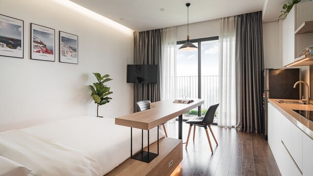 A modern hotel room with a bed, a wooden desk, a dining area, a TV, and a kitchen area. - Mira Housing- Vinhomes D'Capitale  (Hanoi)