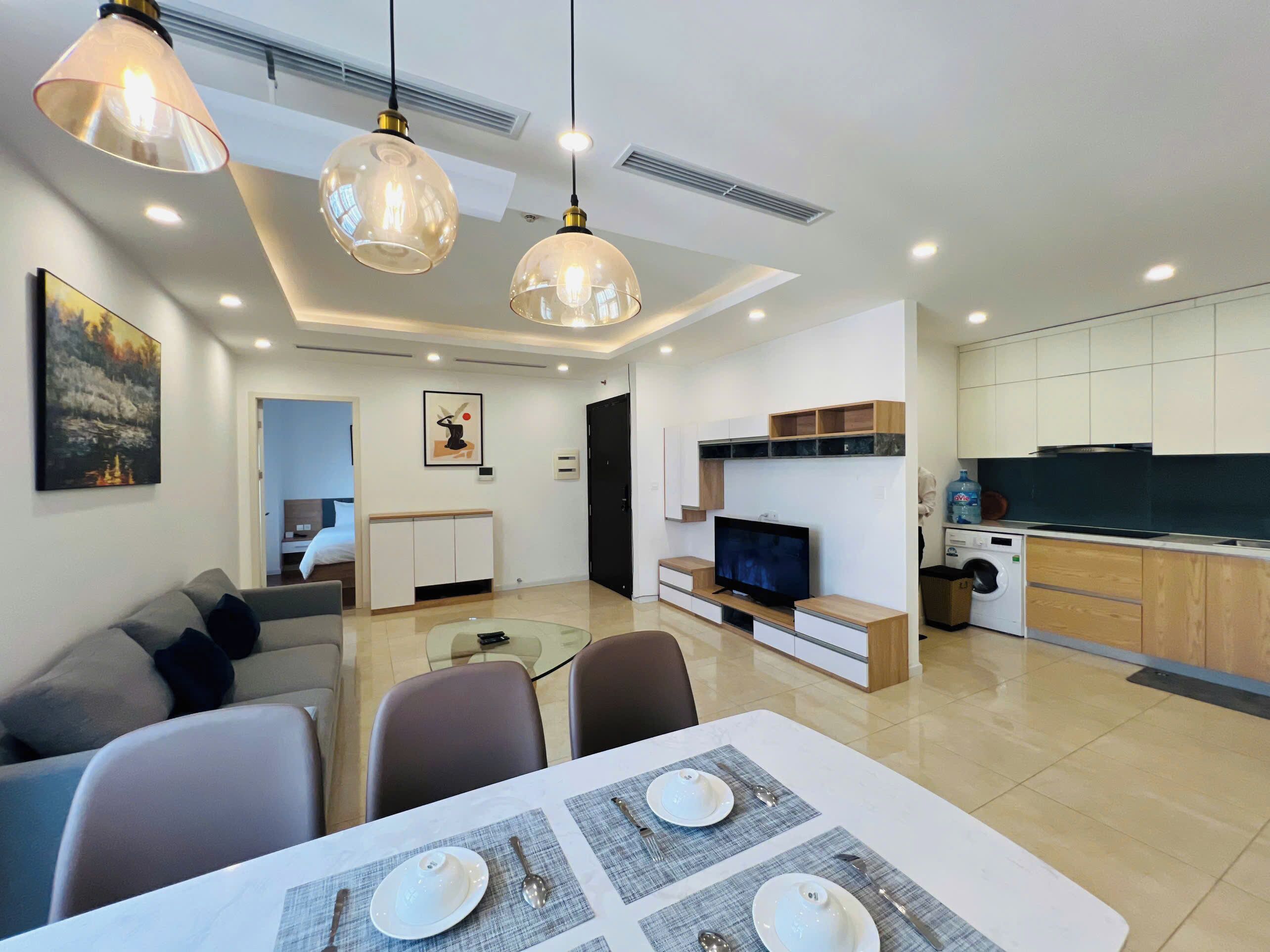 A modern living room with a sofa, dining table set for two, and a TV. There is a kitchen area with cabinets and a washing machine.