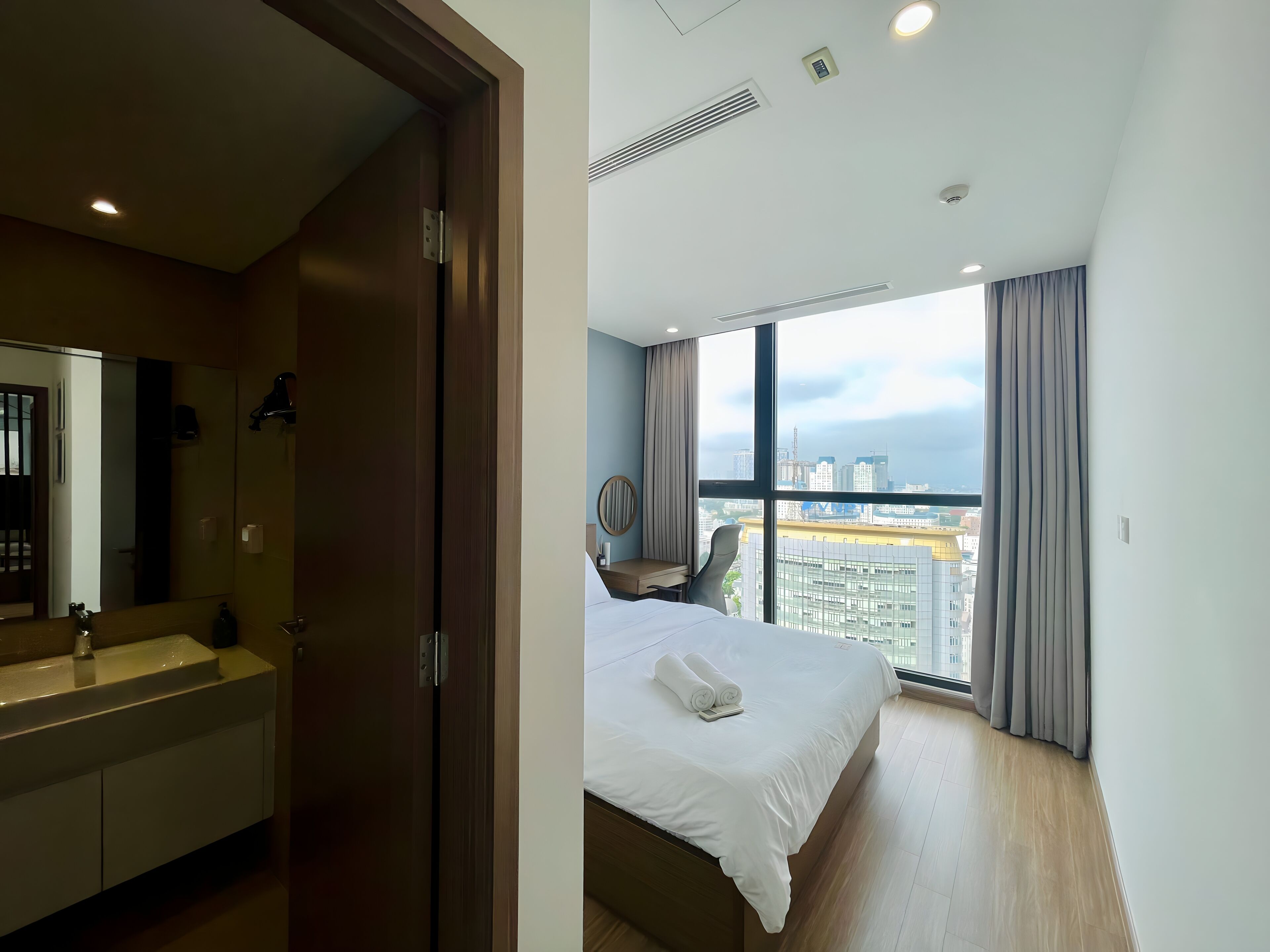 Habitación de hotel con cama, un amplio ventanal con cortinas, baño con espejo y vista panorámica de la ciudad.