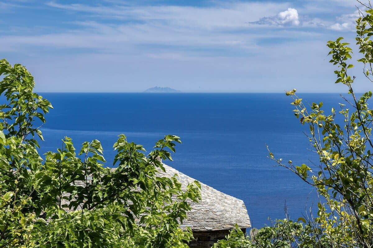 A coastal view with a stone building, lush greenery, and a clear blue sea.