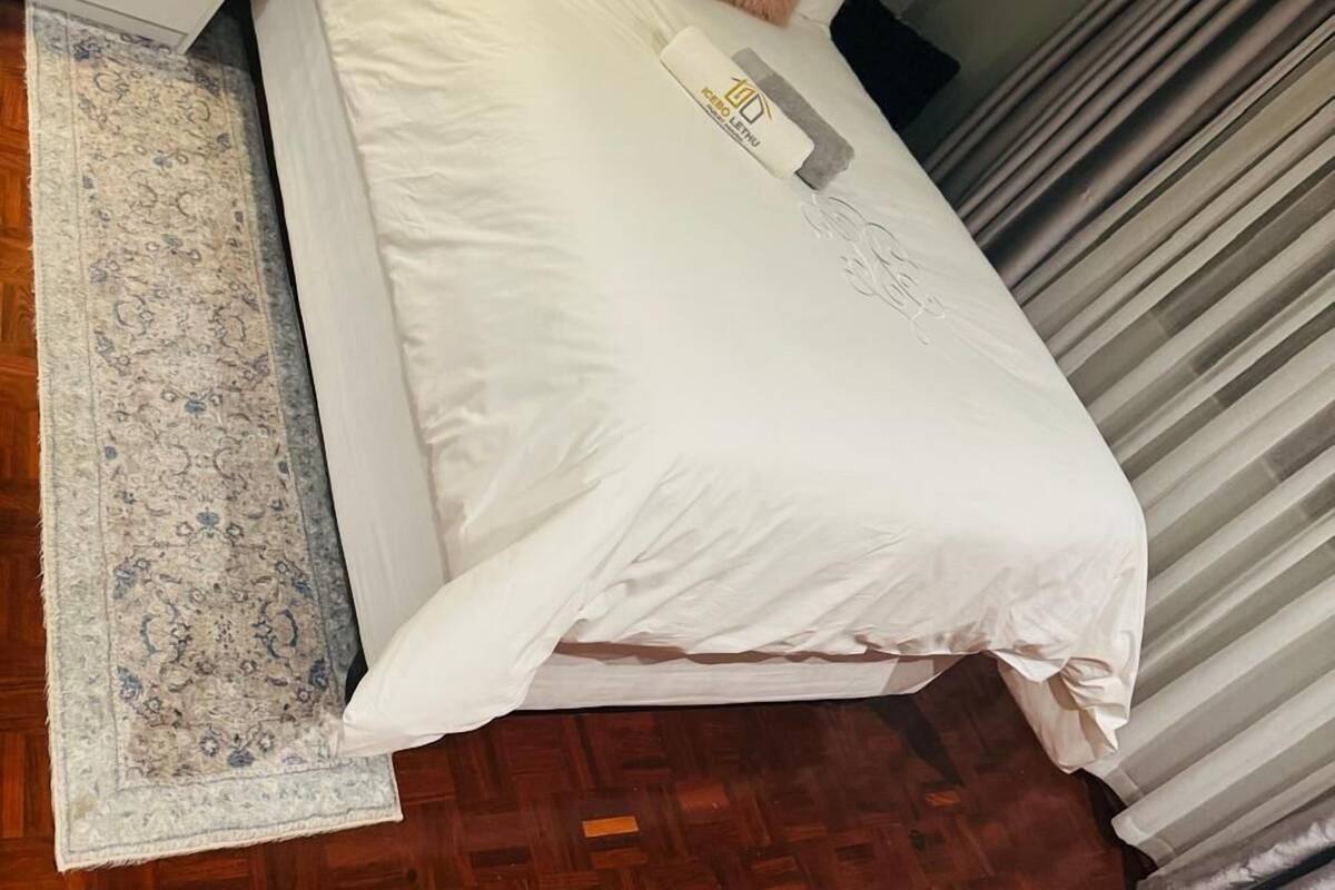 Ein ordentlich bezogenes Bett mit weißen Leinen und einem flauschigen Kopfkissen, ein Nachttisch mit einer Lampe und ein gemustertes Teppich auf Holzdielen.