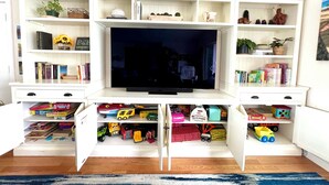 A living room with a TV on a white entertainment unit, surrounded by shelves with books, toys, and decorative items.