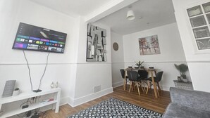 Una sala de estar moderna con un televisor de pantalla plana montado en la pared, un comedor con una mesa y sillas de madera, y un sofá gris.