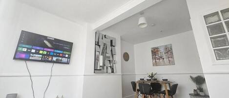 A modern living room with a flat-screen TV mounted on the wall, a dining area with a wooden table and chairs, and a grey sofa.