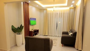 A modern living room with a sofa, a large potted plant, a flat-screen TV, and a window with curtains. - The Residence Ug (Kampala)