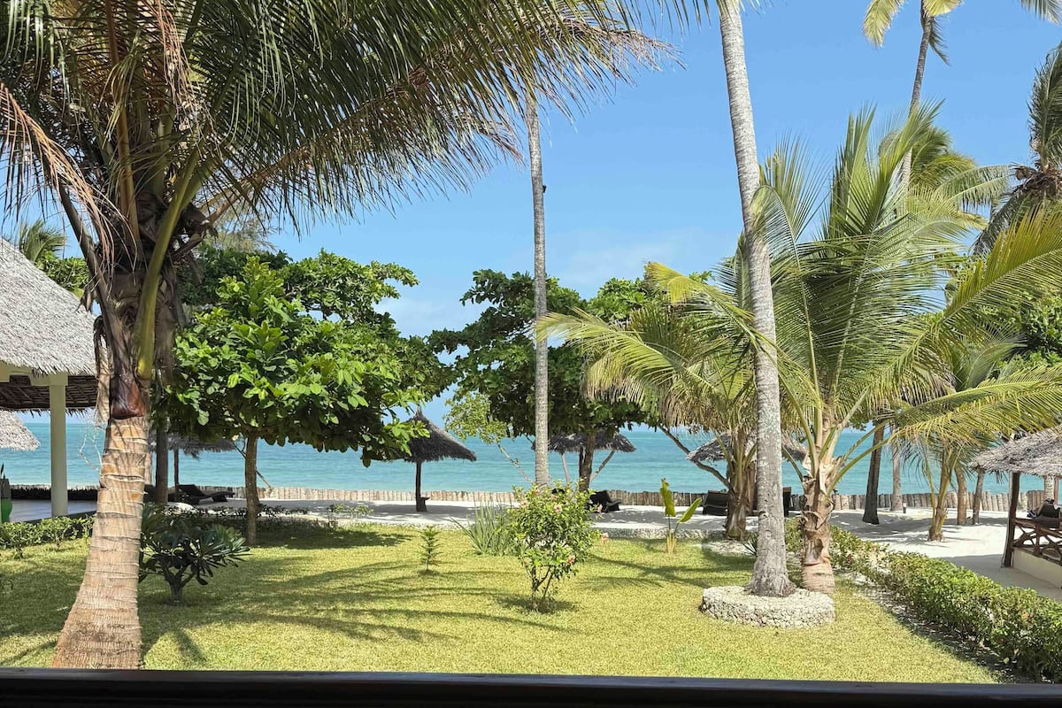 A tropical resort with palm trees, a thatched-roof structure, and a view of the sea.