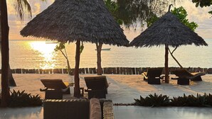 A beachside lounge area with thatched umbrellas, lounge chairs, and a wooden fence overlooking the sea. - MANOLO Beach Resort (Uroa)