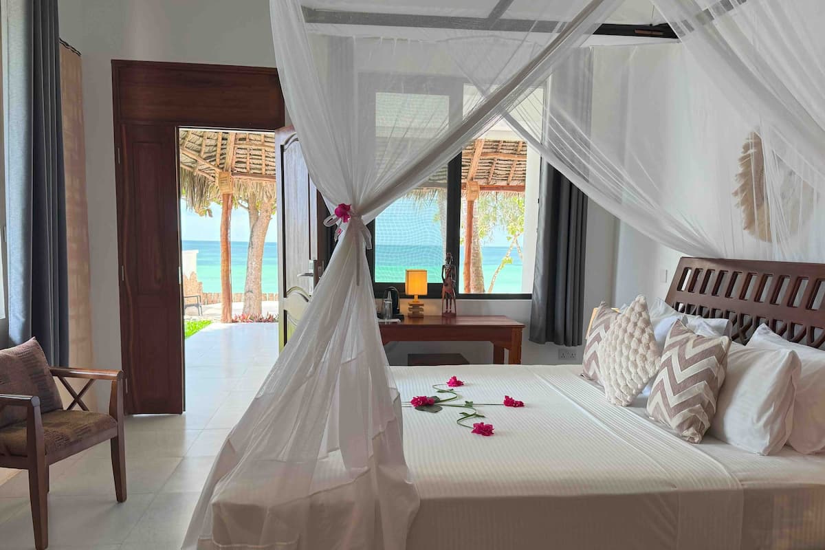 A bedroom with a bed, a nightstand, a chair, and a view of the beach through large windows.