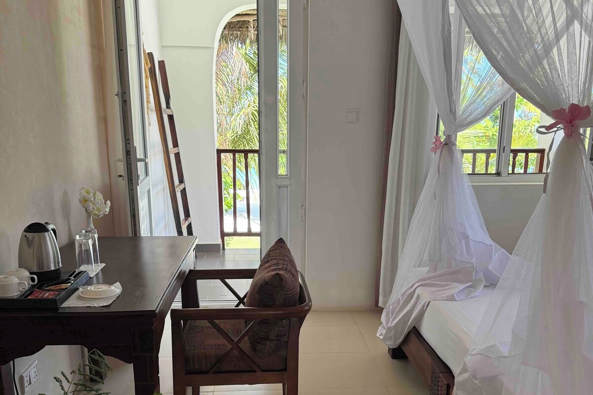 Une chambre avec un lit surmonté d’un dais blanc, une table à manger avec une bouilloire et des tasses, et une chaise avec un coussin.