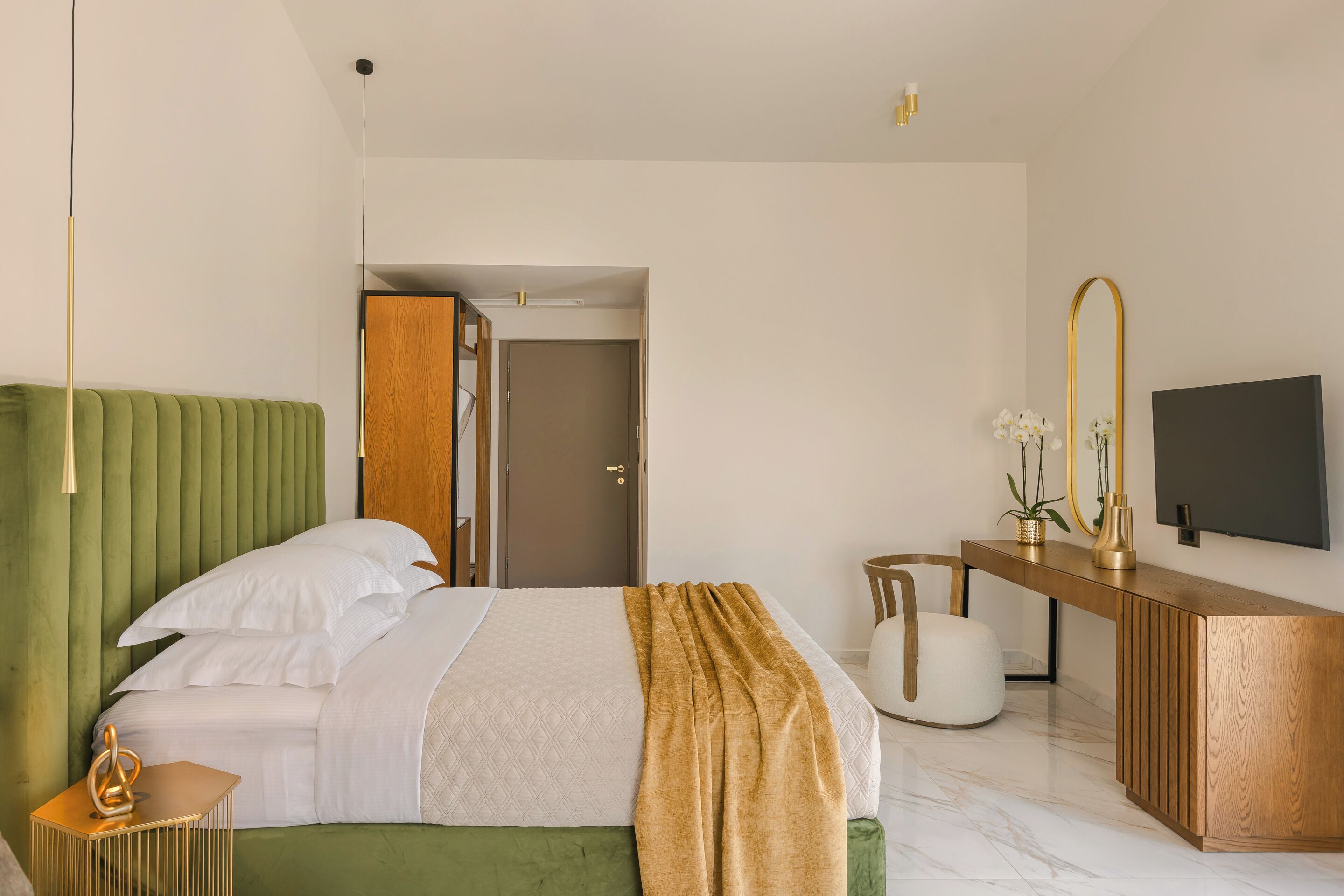 Une chambre d’hôtel moderne avec une tête de lit recouverte d’un tissu vert, un bureau en bois avec un téléviseur à écran plat, une chaise blanche et une petite table d’appoint agrémentée d’un objet décoratif.