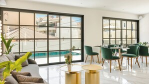 Una habitación de hotel moderna con un ventanal grande, vistas a la piscina, un sofá y un comedor con sillas verdes.