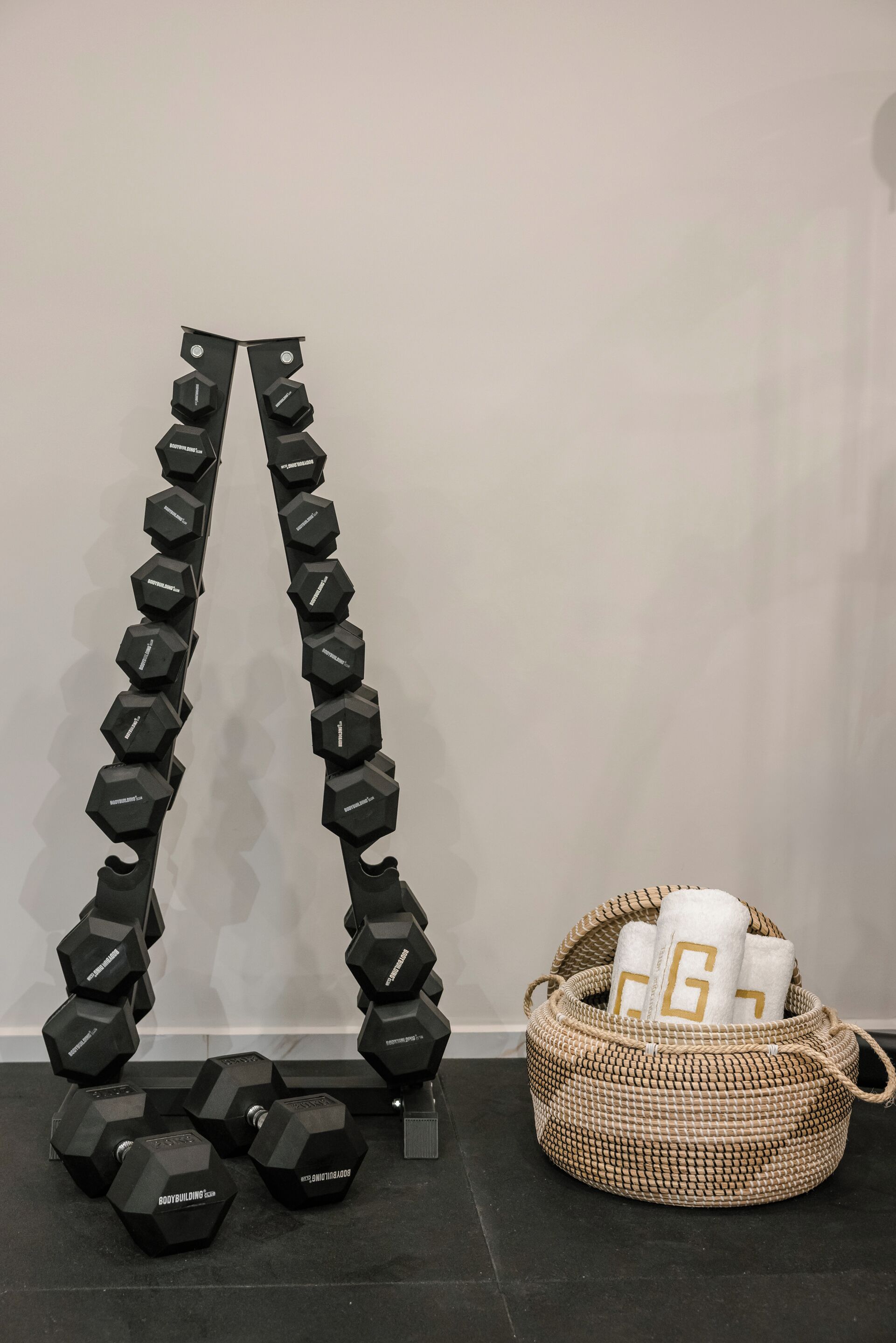 A set of hexagonal dumbbells arranged in a V-shape on a black floor, with a woven basket containing towels with a 'G' logo next to it.