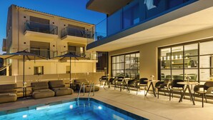 Un área de piscina moderna al aire libre con sillones de descanso y mesas, rodeada por un edificio con balcones y ventanas amplias.