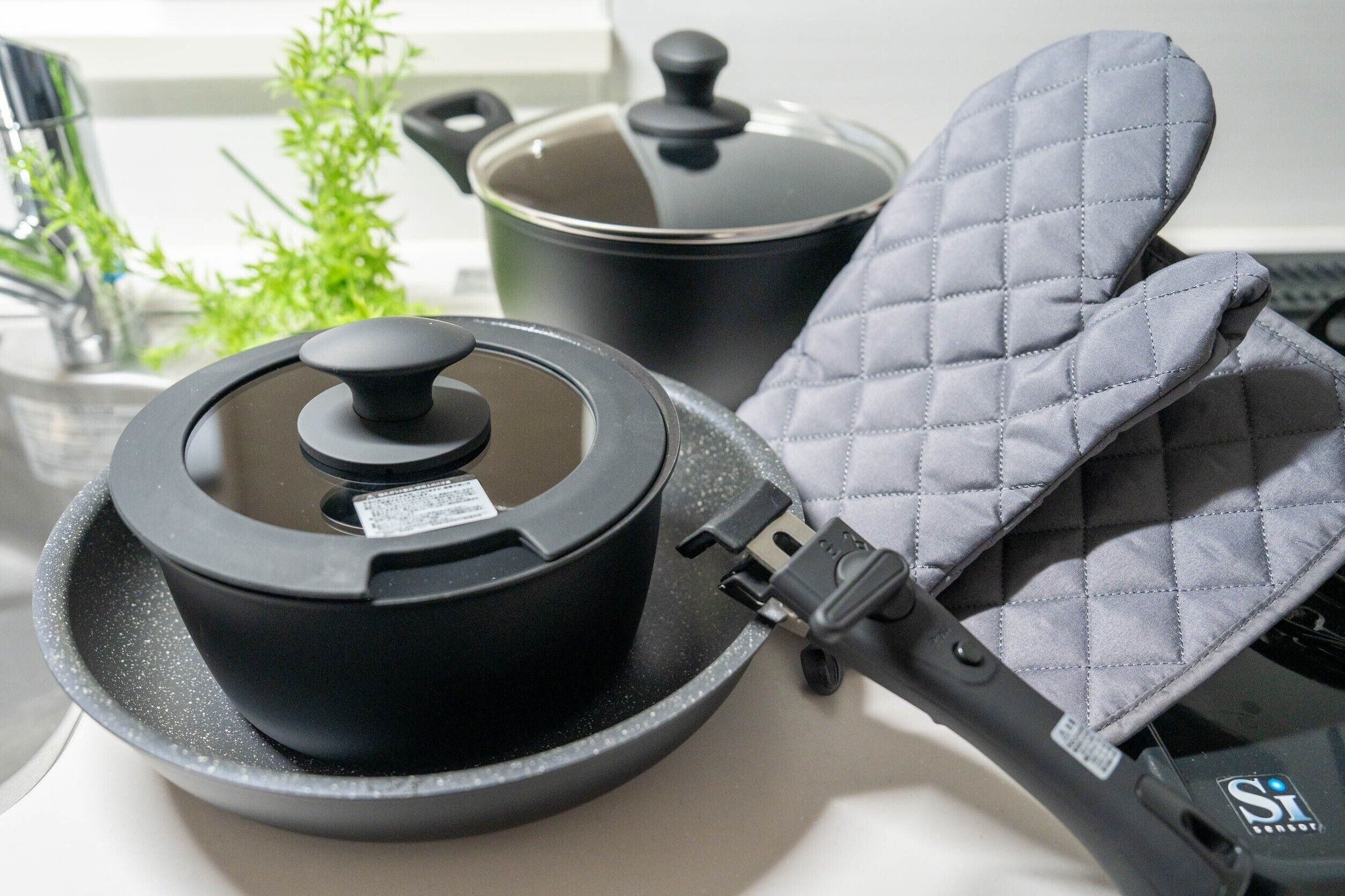 A kitchen with a black electric rice cooker, a pot with a lid, and a pair of gray oven mitts on a countertop.