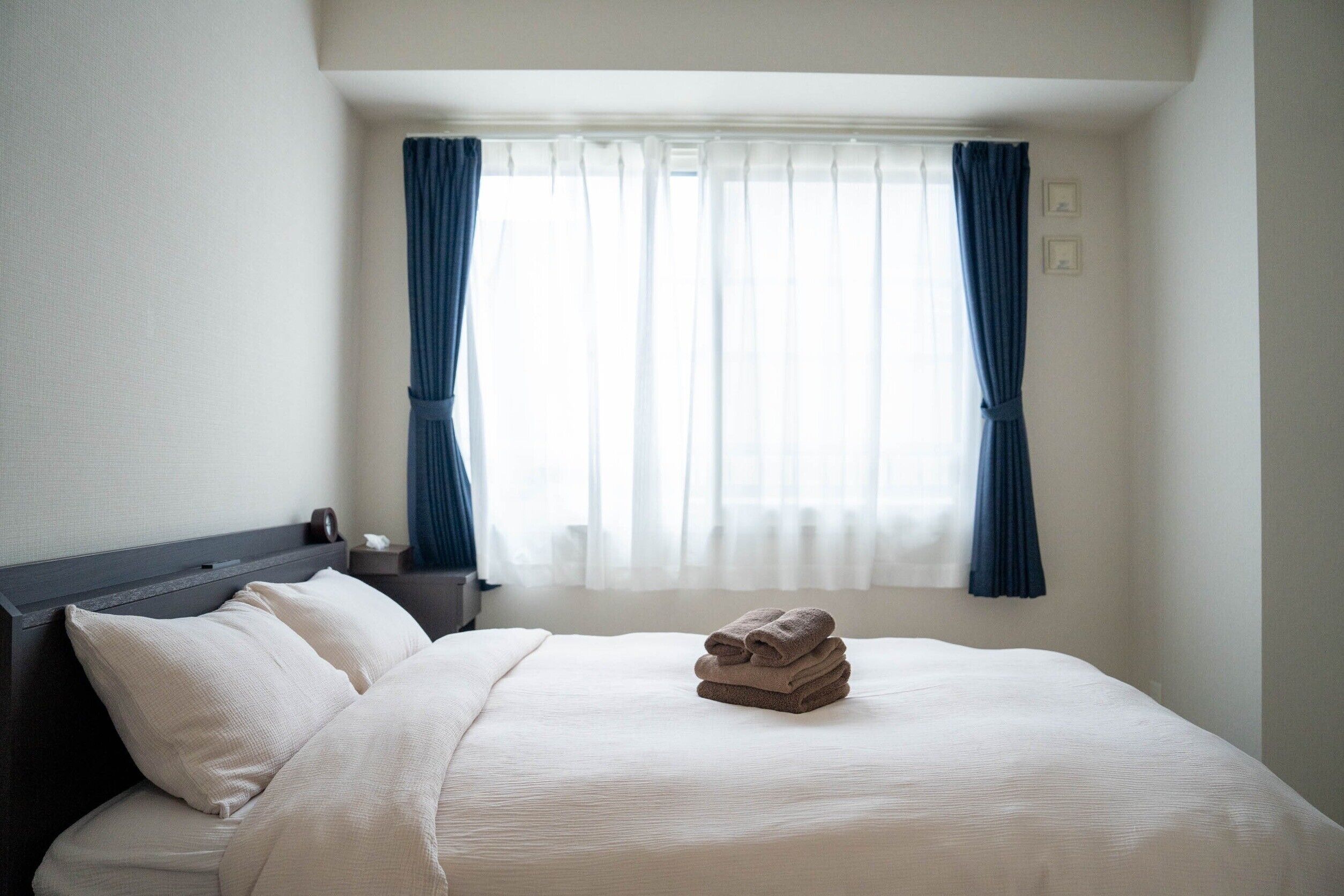 Una cama bien hecha con una toalla doblada, una mesita de noche con lámpara y una ventana con cortinas.