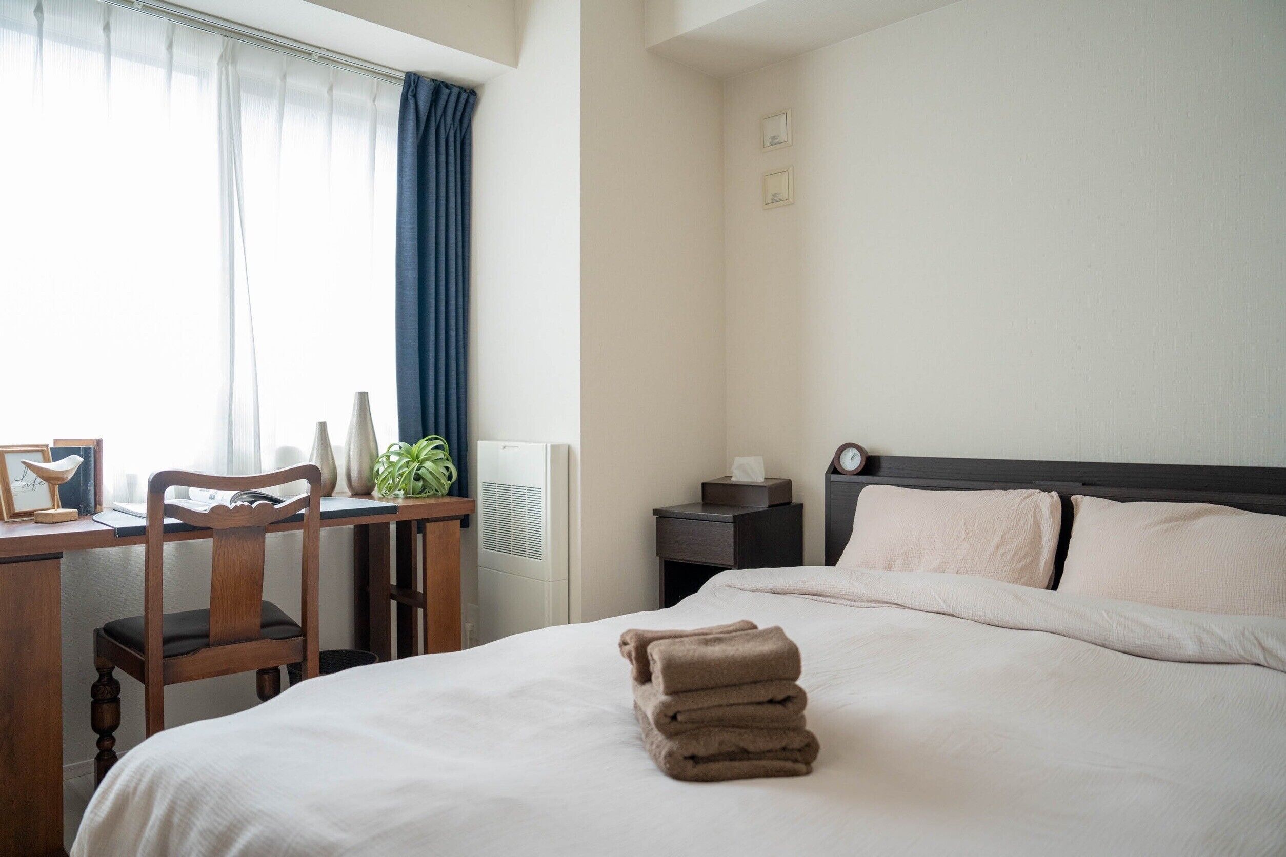 A hotel room with a bed, a desk with a chair, a small table with a plant, and a radiator.