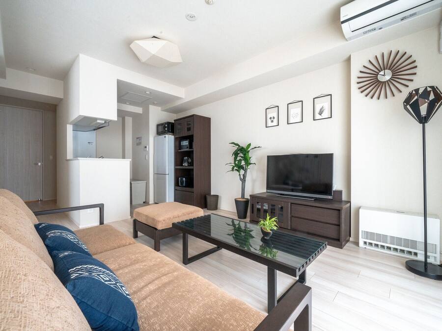 A modern living room with a sofa, coffee table, and a flat-screen TV. There is a kitchen area in the background with cabinets and a microwave.