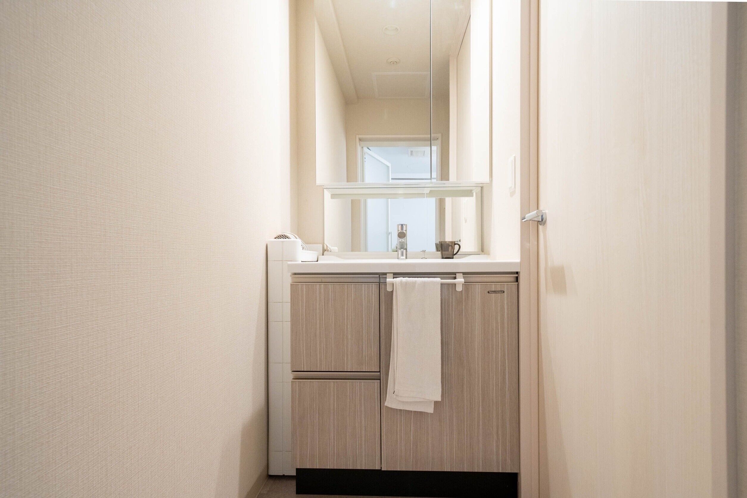Un couloir étroit mène à une salle de bain équipée d’un lavabo, d’un miroir et d’une serviette accrochée à la porte.