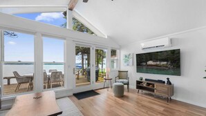 Un salon spacieux doté d’un grand téléviseur, d’un plancher en bois et offrant une vue sur la mer grâce à de grandes portes vitrées.