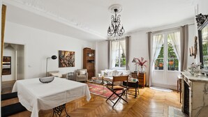 Een ruime kamer met een houten visgraatvloer, een wit bed, een eettafel, een kroonluchter en uitzicht op groen.