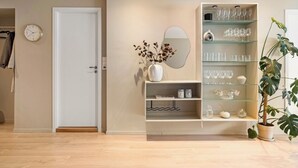 Habitació amb sòl de fusta, una porta blanca, un rellotge de paret, un mirall, una prestatgeria de vidre amb recipient de vidre, un vas amb flors seques i una planta en test.