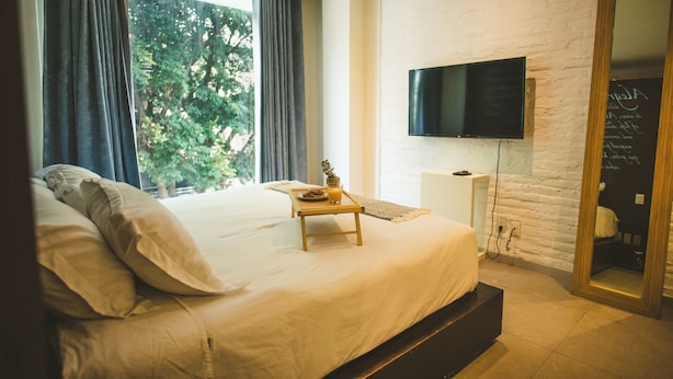 Une chambre à coucher avec un lit, une table de chevet sur laquelle se trouve un plateau, un téléviseur fixé au mur en briques, et une grande fenêtre avec des rideaux.