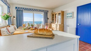 Une cuisine dotée d’un grand îlot central, un coin repas offrant une vue sur la mer et une porte bleue.