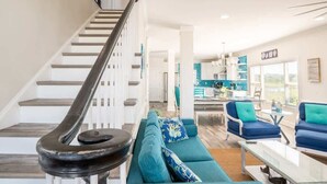 A modern living room with a staircase, teal sofas, and a kitchen in the background. - The Saltwater Shack: Early Bird Special! (Oak Island)