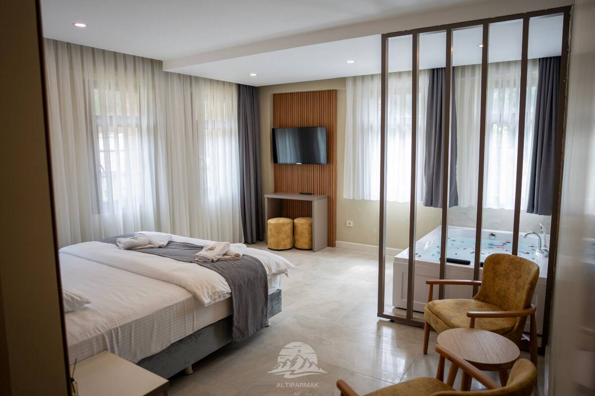A modern hotel room with a large bed, a sitting area with a chair and ottoman, a TV mounted on the wall, and a bathtub with a shower.