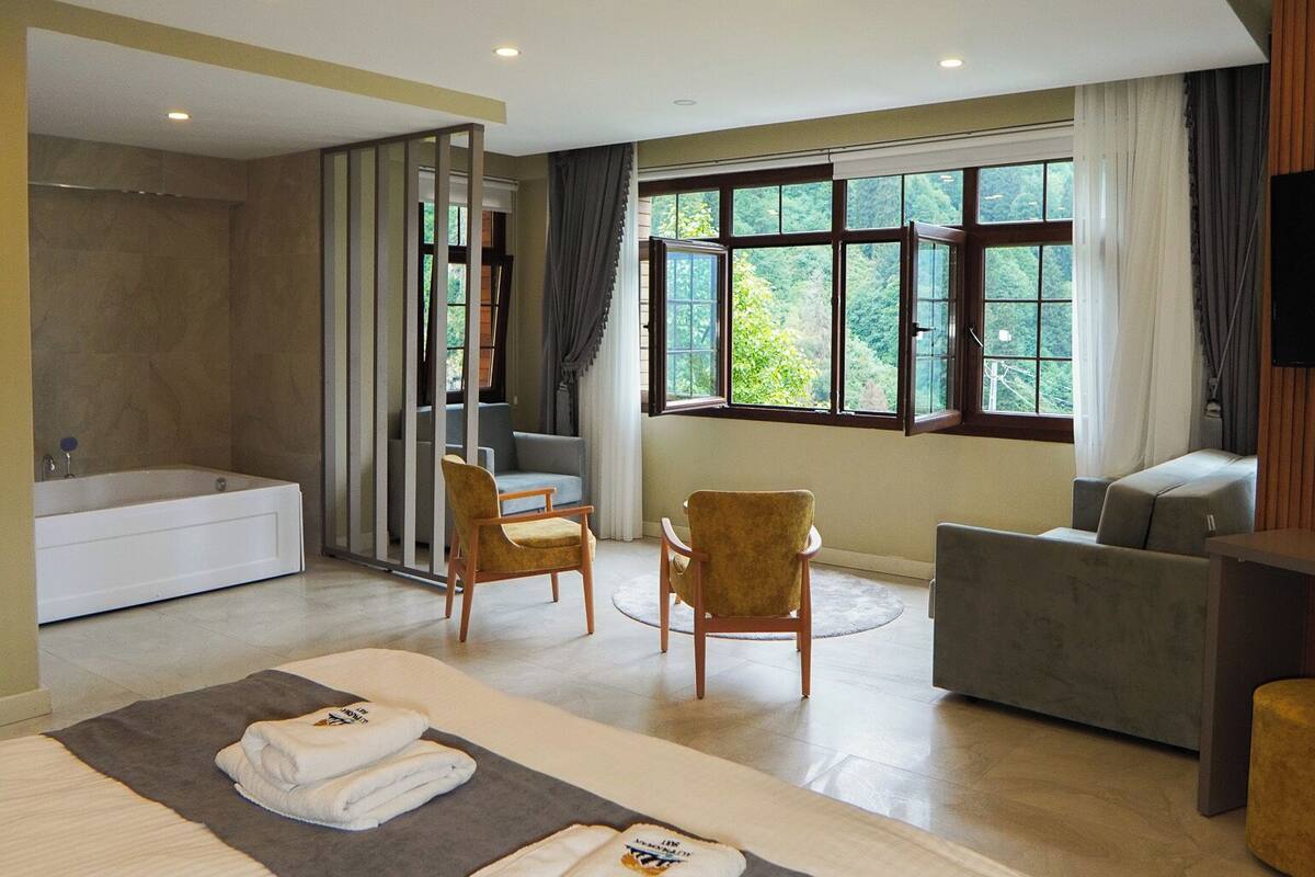 A modern hotel room with a large bed, a bathtub, two armchairs, a sofa, and a TV.