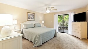 Ein Schlafzimmer mit einem Bett, einem Deckenventilator, einem Fernseher, einer Kommode und einem Fenster mit Vorhängen.