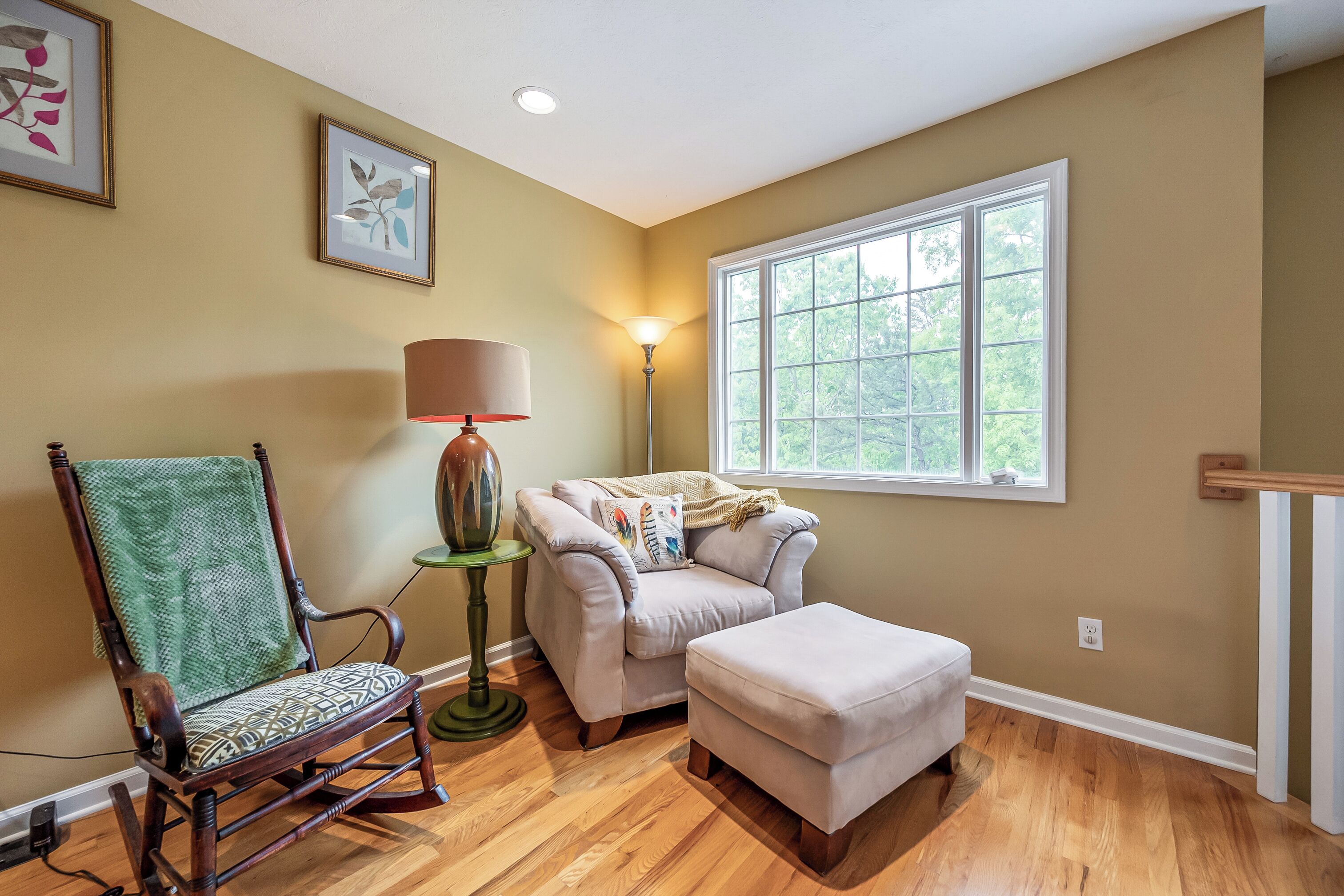 A room with a rocking chair, a sofa, and an ottoman. There is a window with a view of trees and a lamp on a side table.