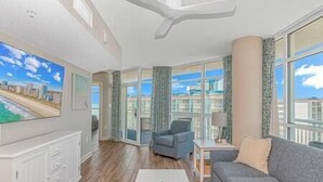 A modern living room with a grey sofa, a coffee table, and a city view through large windows. - 2BR Penthouse End Unit w/ Indoor Waterparks! (Myrtle Beach)
