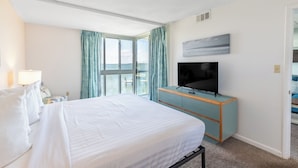 A hotel room with a large bed, a TV on a light blue cabinet, and a view of the sea through a large window with curtains. - 2BR Near Beach | Bay Views + Jolly Roger Fun (Ocean City)
