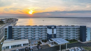 A multi-story hotel with a flat roof, a parking area, and a view of the water and sunset. - 2BR Near Beach | Bay Views + Jolly Roger Fun (Ocean City)